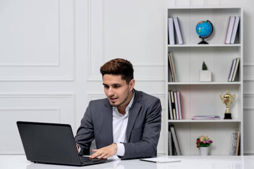 A professional man in a suit works on a laptop at a modern office desk, with bookshelves and a globe in the background.