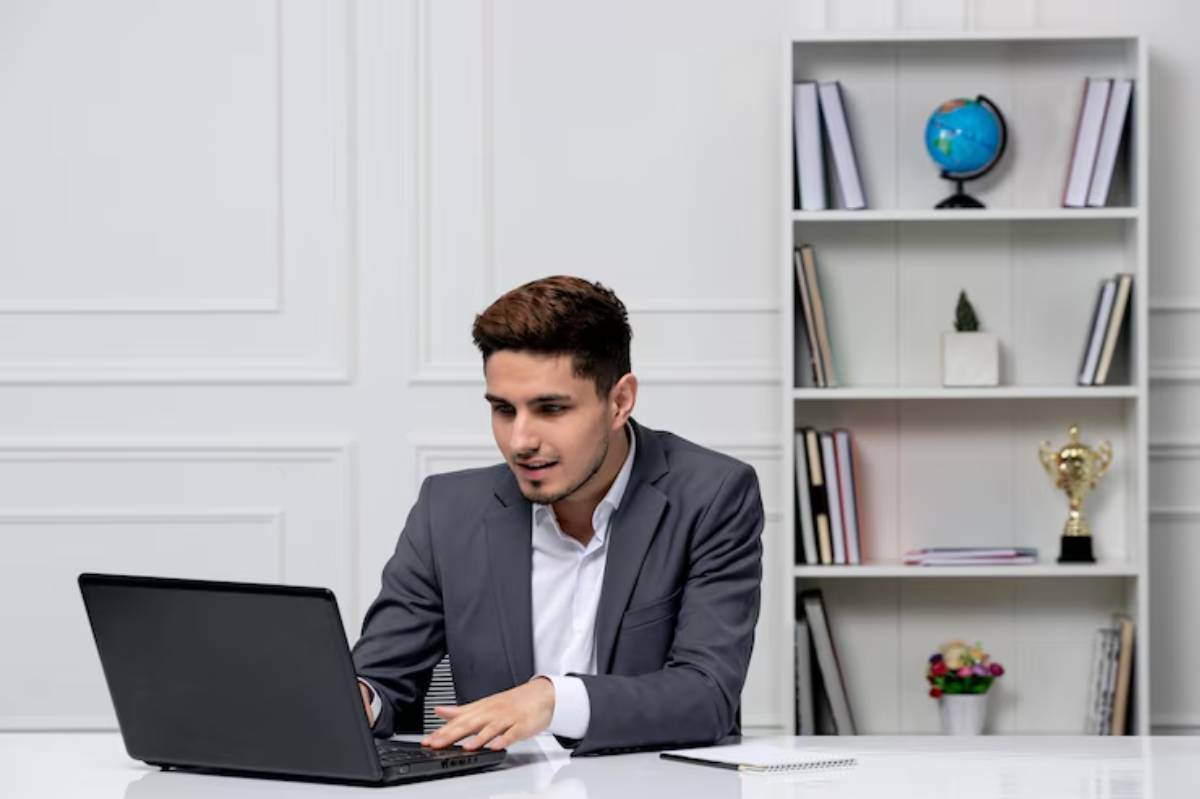 A professional man in a suit works on a laptop at a modern office desk, with bookshelves and a globe in the background.