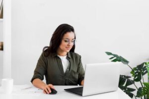 A person in a green shirt uses a laptop at a white desk, surrounded by plants and office supplies.