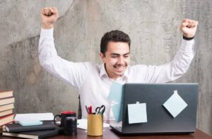 A person in a white shirt celebrates triumphantly at a desk cluttered with books, stationery, and a laptop displaying sticky notes.