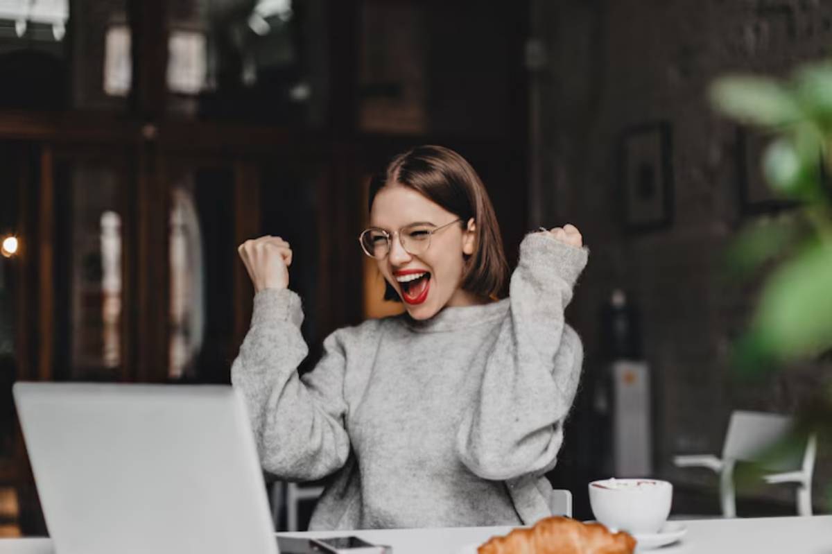A person in a cozy gray sweater celebrates with raised fists at a table with a laptop, a croissant, and a cup of coffee.
