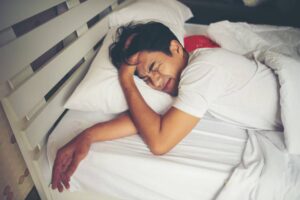 A person lying on a bed with white linens, resting their head on their arm, surrounded by soft pillows.