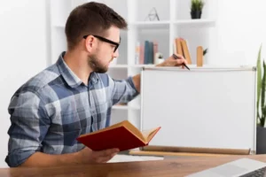 A person in a plaid shirt holds an open book while writing on a blank whiteboard in a well-lit, organized workspace.