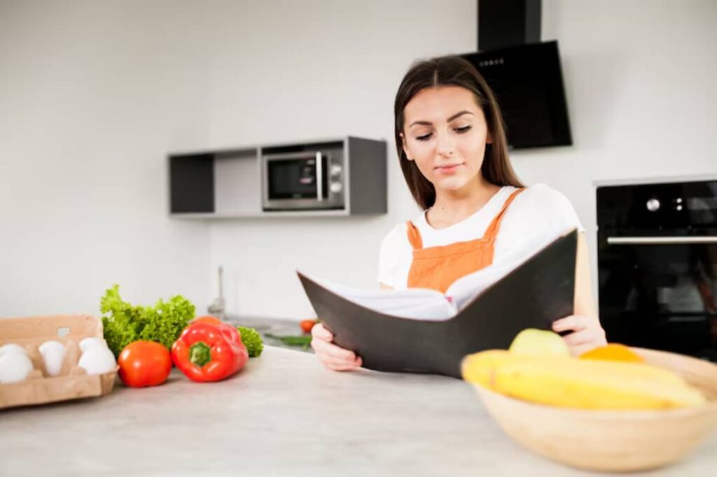 A person in an orange apron reading a cookbook in a modern kitchen with fresh vegetables and fruit on the counter.