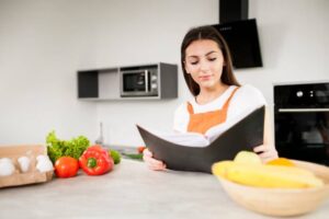 A person in an orange apron reading a cookbook in a modern kitchen with fresh vegetables and fruit on the counter.