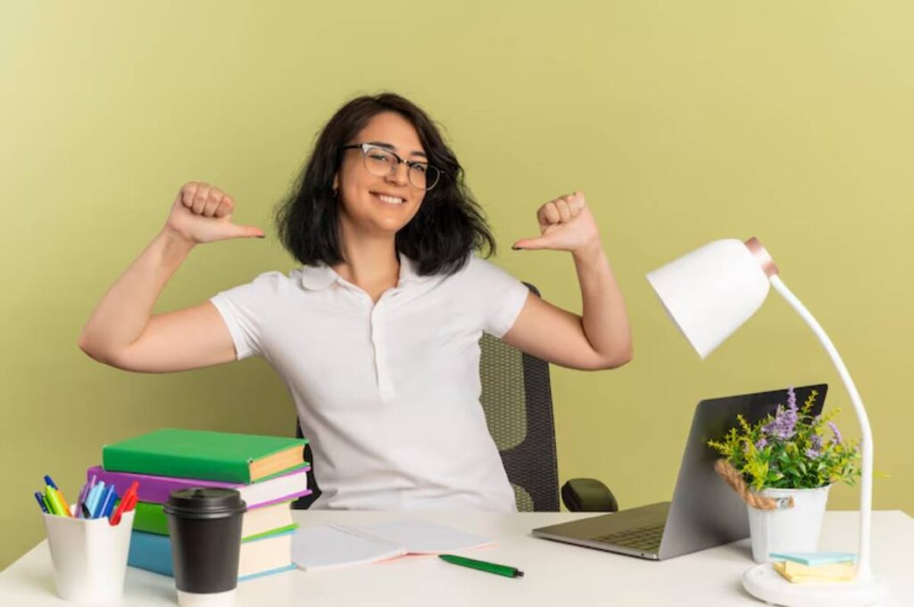A person in a white shirt cheerfully gives a thumbs-up at a desk with books, a laptop, stationery, and a lamp against a light green wall.