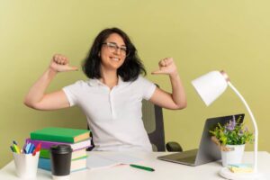 A person in a white shirt cheerfully gives a thumbs-up at a desk with books, a laptop, stationery, and a lamp against a light green wall.
