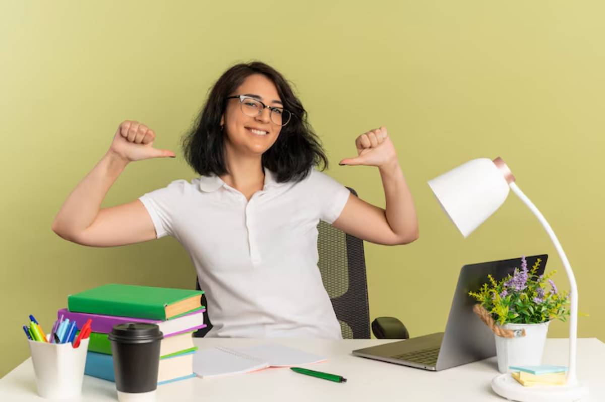 A person in a white shirt cheerfully gives a thumbs-up at a desk with books, a laptop, stationery, and a lamp against a light green wall.