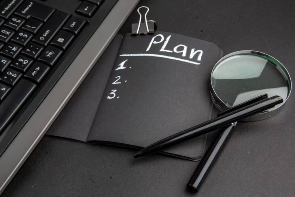 A black notebook labeled Plan with numbered lines, a magnifying glass, and pens, beside a computer keyboard on a dark surface.