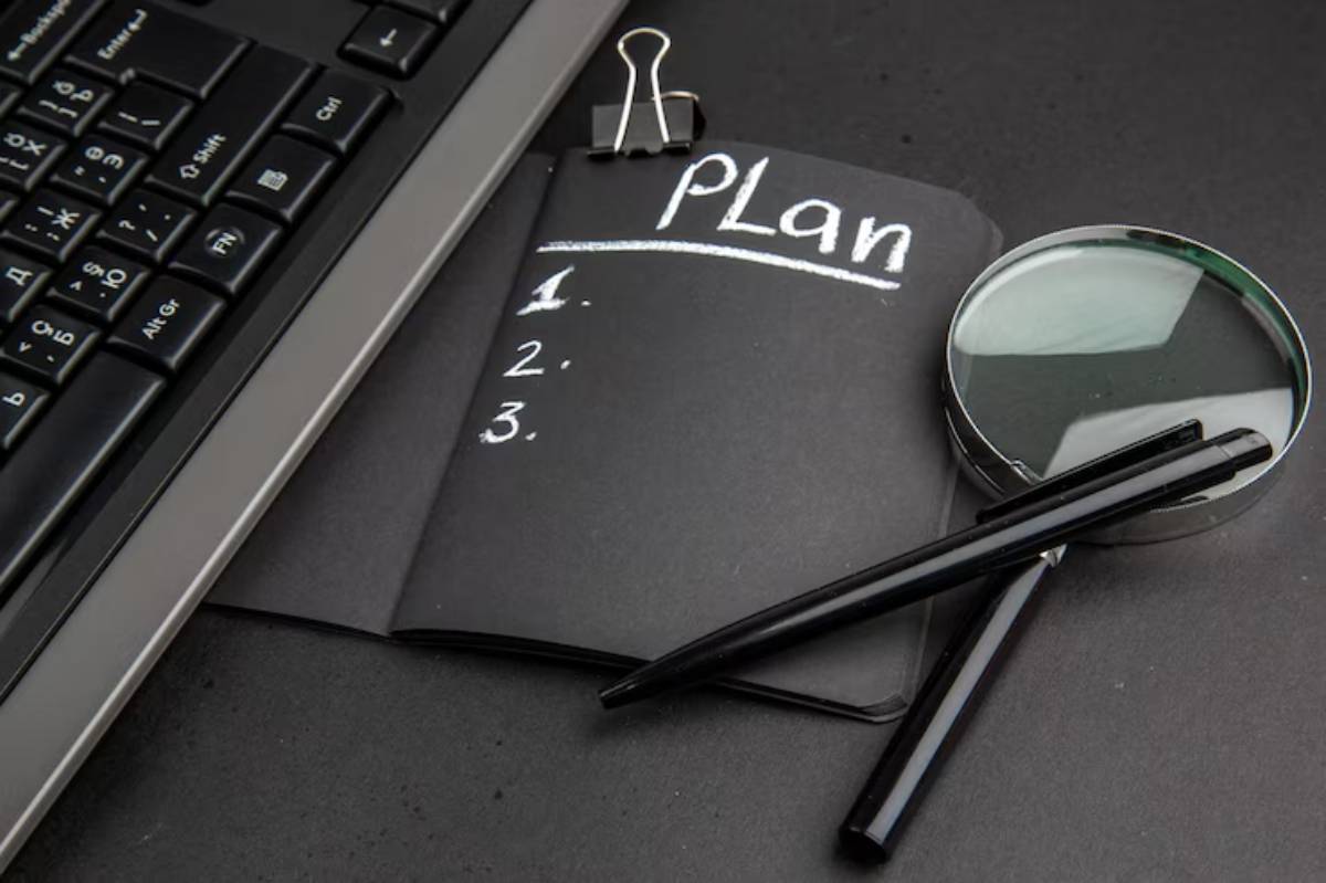 A black notebook labeled Plan with numbered lines, a magnifying glass, and pens, beside a computer keyboard on a dark surface.