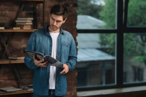 A person in a denim shirt holds a magazine, standing near a large window with greenery visible outside. Books are stacked on a nearby shelf.