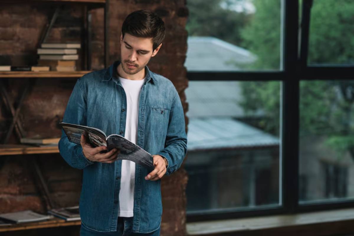 A person in a denim shirt holds a magazine, standing near a large window with greenery visible outside. Books are stacked on a nearby shelf.