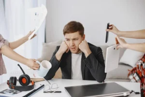 A stressed individual sits at a desk, overwhelmed by papers, a mug, a tablet, and a phone, amidst a cluttered workspace.