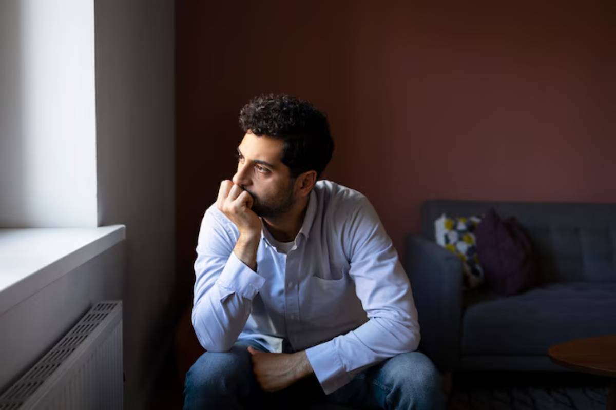 A person sits pensively on the floor, wearing a light blue shirt, in a cozy room with a gray couch and natural light coming through a window. 