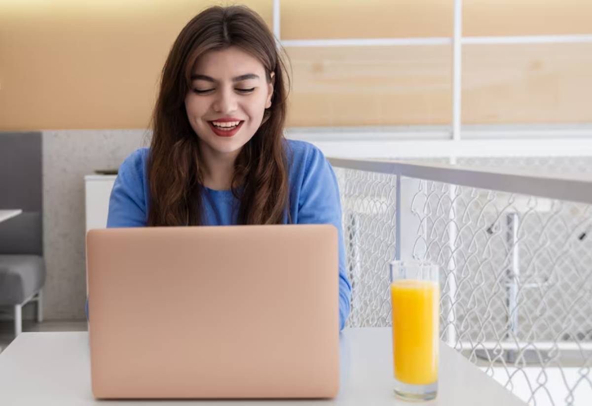 A person in a blue sweater is seated at a table with a laptop and a glass of orange juice in a modern café setting. 