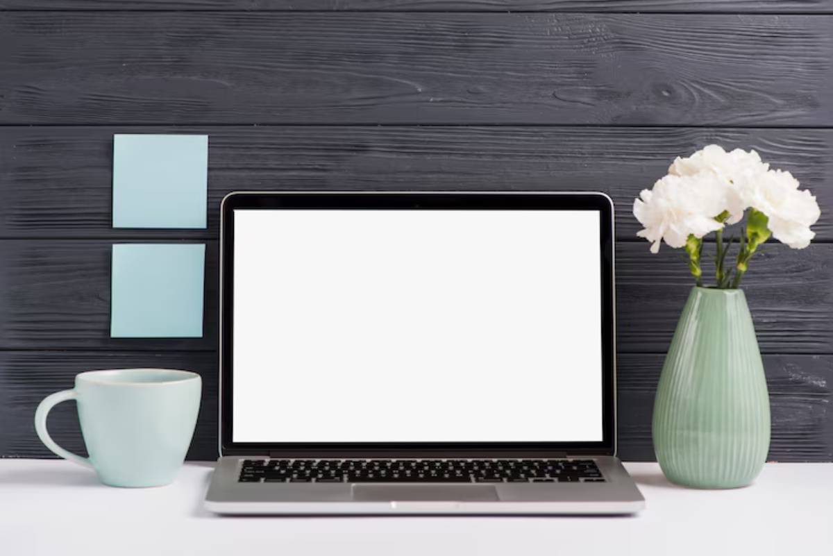 A minimal workspace featuring a laptop with a blank screen, a mint coffee mug, two sticky notes, and a green vase with white flowers.