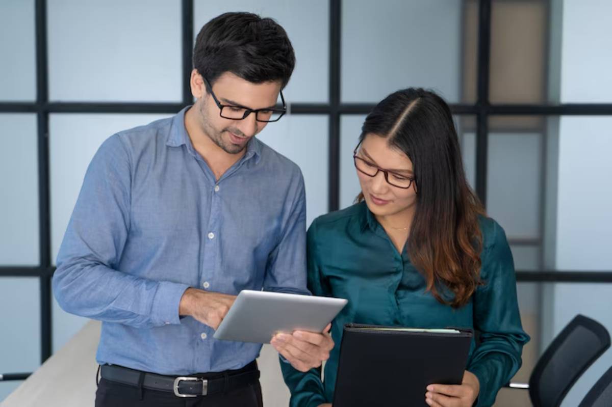 Two professionals are discussing data on a tablet in a modern office setting, surrounded by large windows and a conference table.