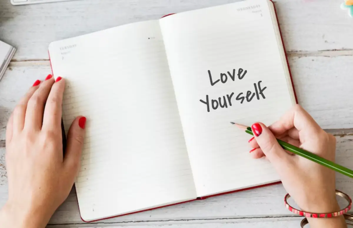 A hand with red nail polish writes Love Yourself in an open notebook with a green pencil on a wooden table. 