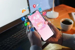 A person holds a smartphone displaying floating social media icons while a laptop and coffee cup sit on a desk in a dimly lit room.