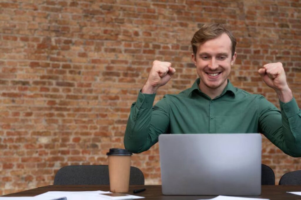 A man in a green shirt raises his fists in celebration, seated at a desk with a laptop, coffee cup, and documents, against a brick wall.