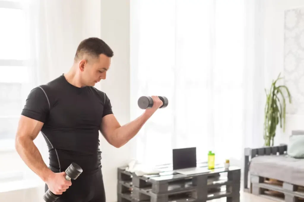 A man in a black workout shirt lifts a dumbbell in a bright room with a laptop and a plant.