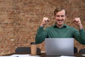 A man in a green shirt raises his fists in celebration, seated at a desk with a laptop, coffee cup, and documents, against a brick wall.
