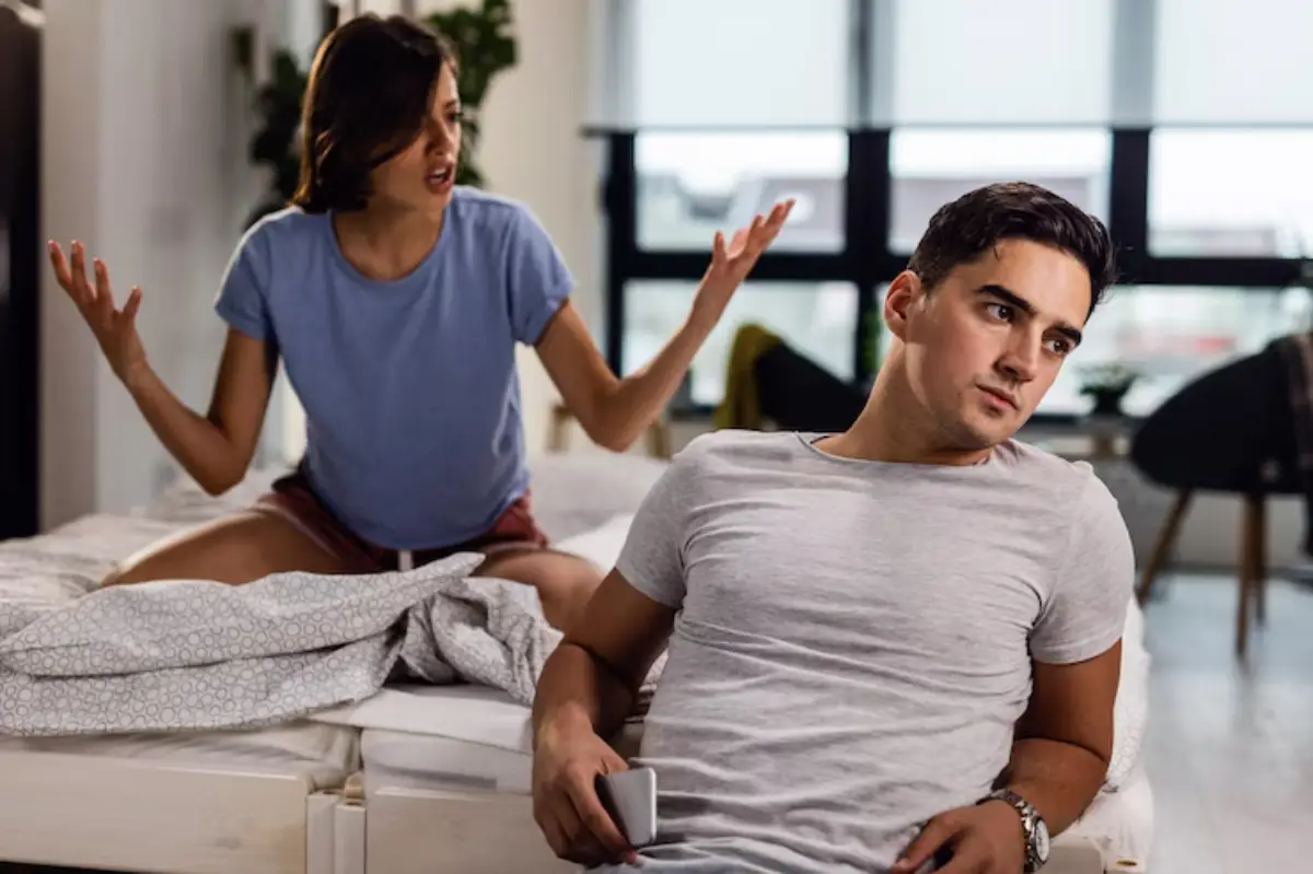 A woman sits on a bed, gesturing expressively, while a man beside her appears uninterested and sits with a phone in hand. 