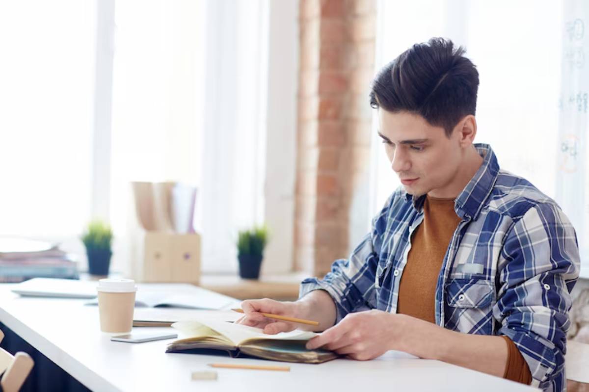 A young man in a plaid shirt studies at a desk, with a notebook and coffee, surrounded by natural light and green plants.