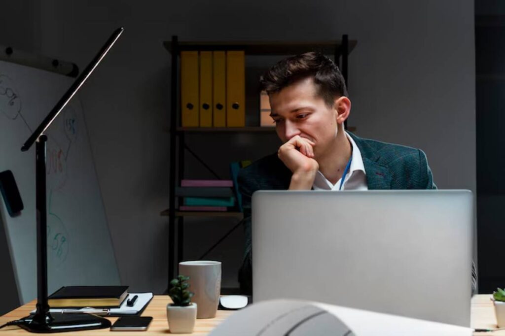 A person sits at a desk, deep in thought, with a laptop, a lamp, a mug, and a small plant on the tabletop in a dimly lit room.