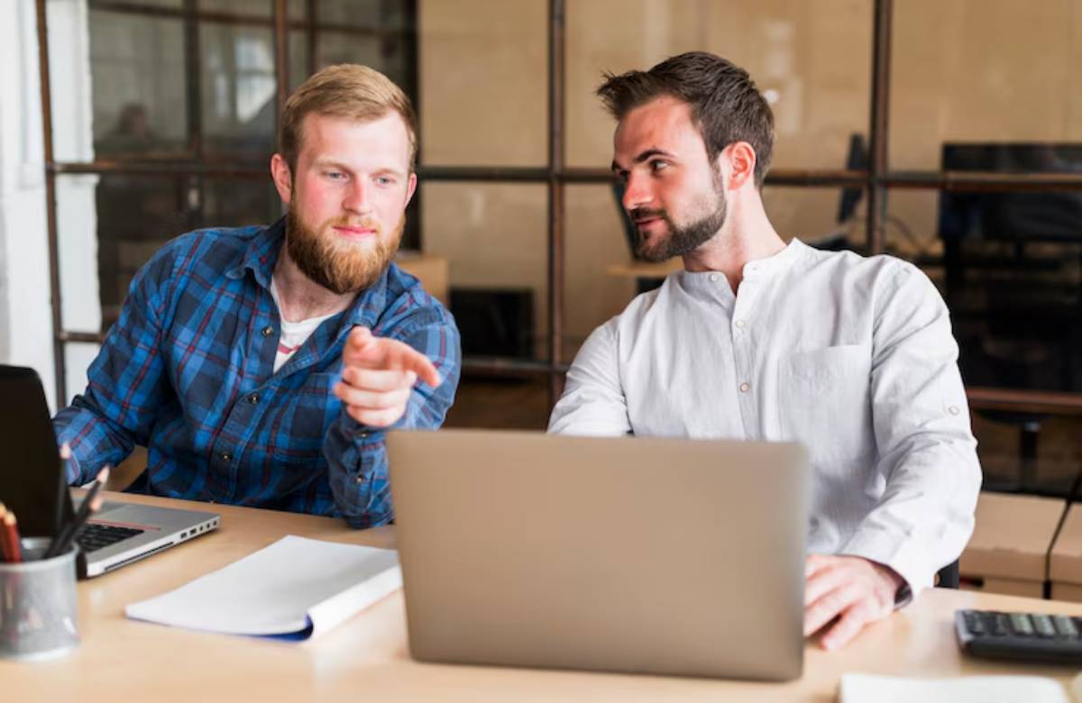 Two men collaborate at a desk with laptops and paperwork, engaged in discussion in a modern office setting.