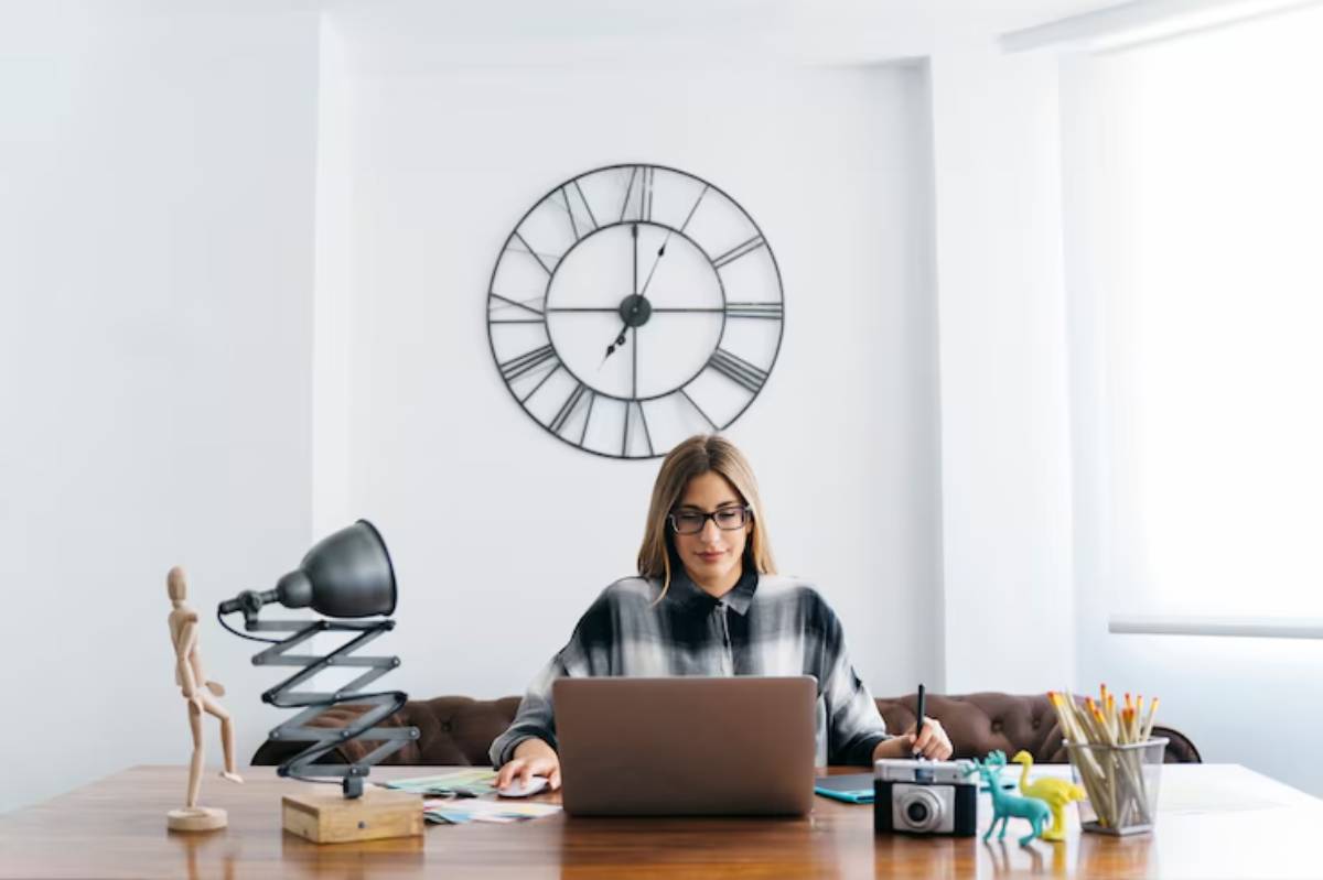 A person sits at a wooden desk with a laptop, lamp, art supplies, a camera, and a clock on the wall behind them. 