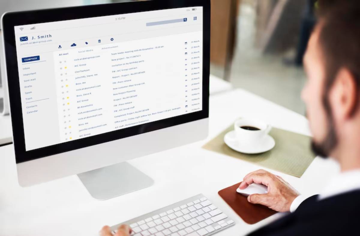 A person views their email inbox on a desktop computer while holding a mouse, with a coffee cup on a table nearby.