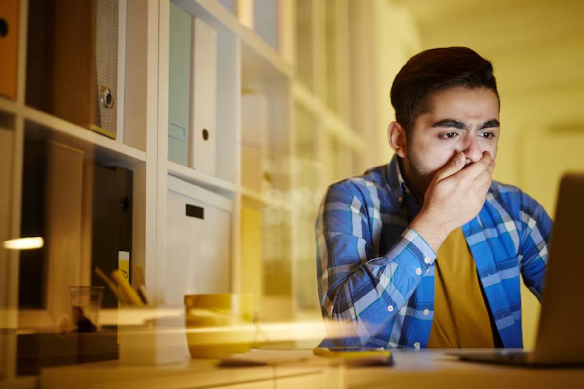 A person in a blue checkered shirt sits at a desk, leaning on their hand, looking thoughtfully at a laptop in a cozy workspace.