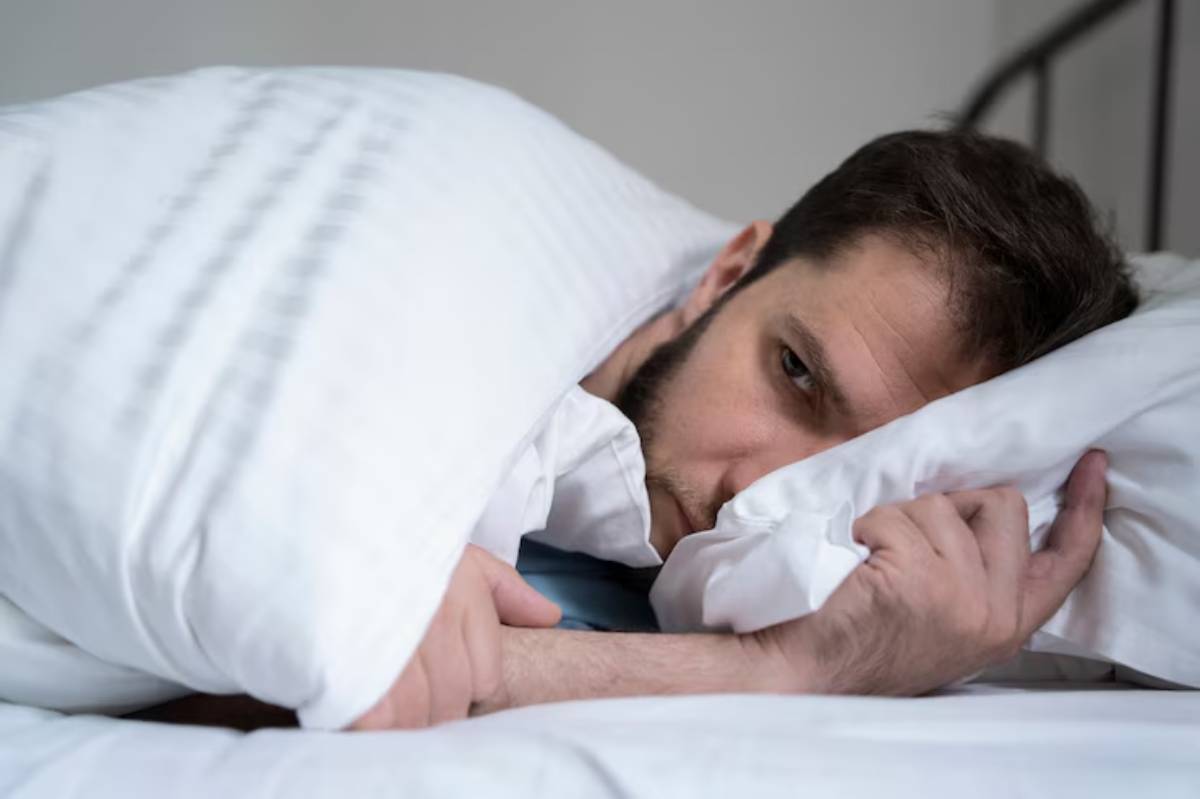 A person lies in bed, covered with a white blanket, their arms tucked closely around themselves, suggesting a feeling of comfort or rest.