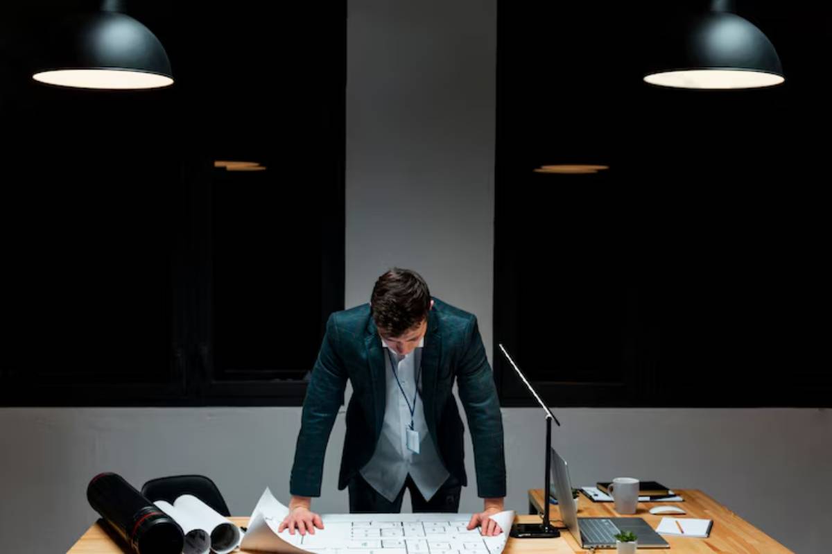 A suited individual leans over blueprints on a desk, surrounded by modern office equipment and under dim lighting. 
