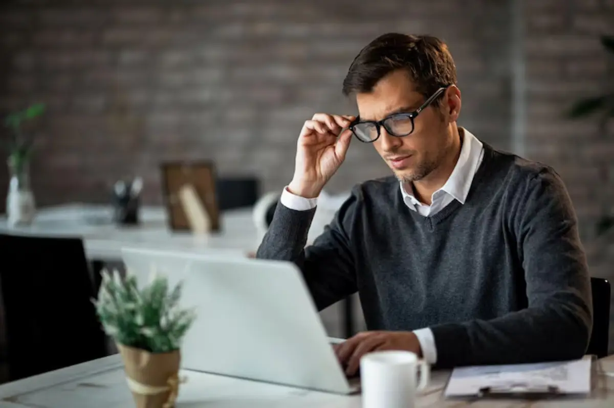 A man in a gray sweater thoughtfully types on a laptop at a stylish desk with plants and office supplies in a cozy setting. 