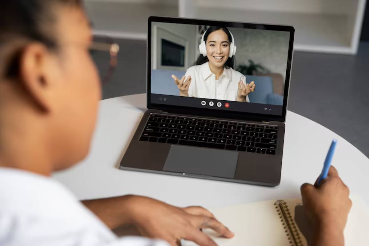 A person seated at a table taking notes while participating in a video call on a laptop.