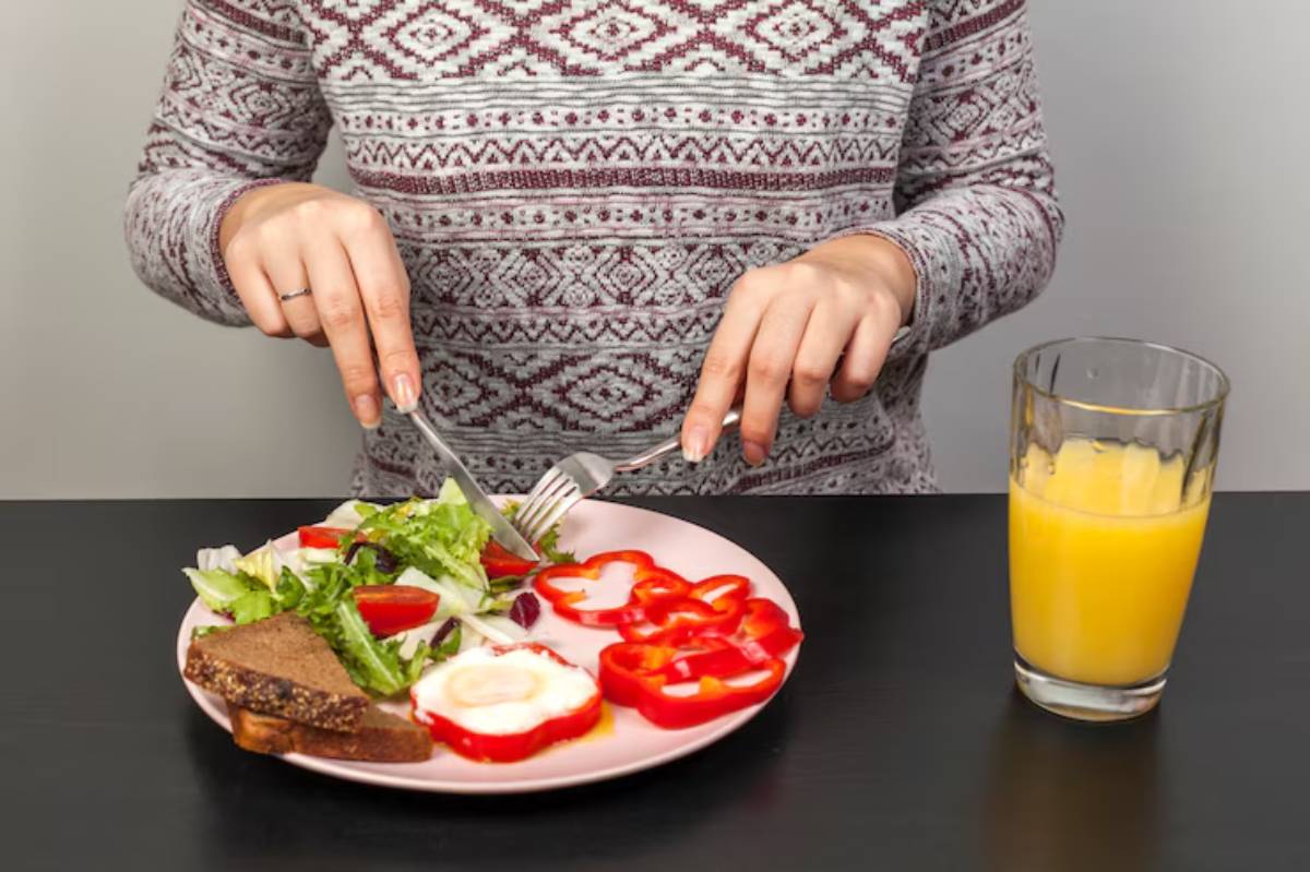A person cuts salad and red peppers on a pink plate while holding a fork and knife, with a glass of orange juice nearby. 