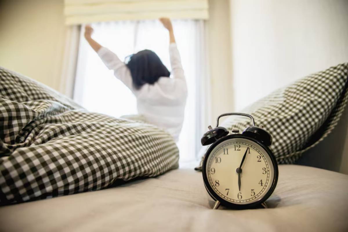 A woman stretches in bed, waking up, with a vintage alarm clock in focus on the bedspread. Sunlight filters through the window.