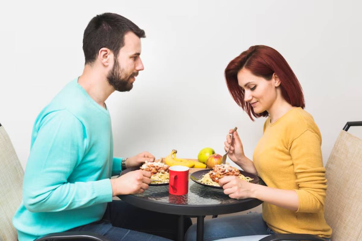 A couple enjoys a meal of pasta and fruit at a round table, with a red mug and bananas prominently displayed.