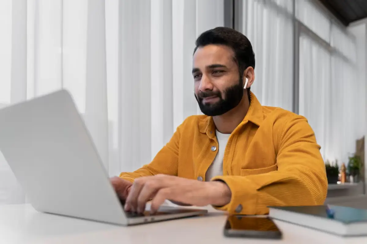 A person wearing wireless earbuds types on a laptop at a bright workspace, with a notebook and smartphone on the table. 
