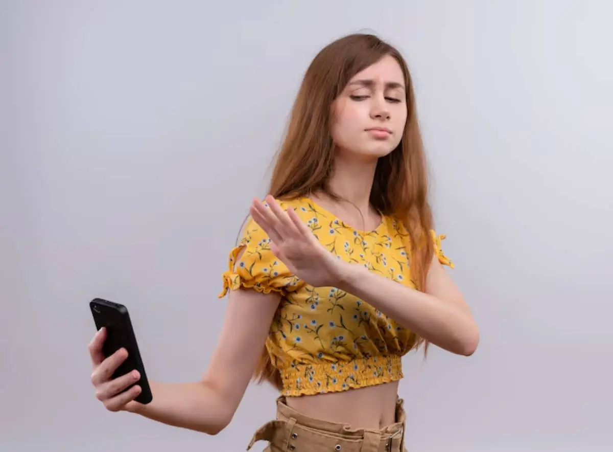 A young woman in a floral yellow crop top holds a phone in one hand while gesturing with the other against a light background. 