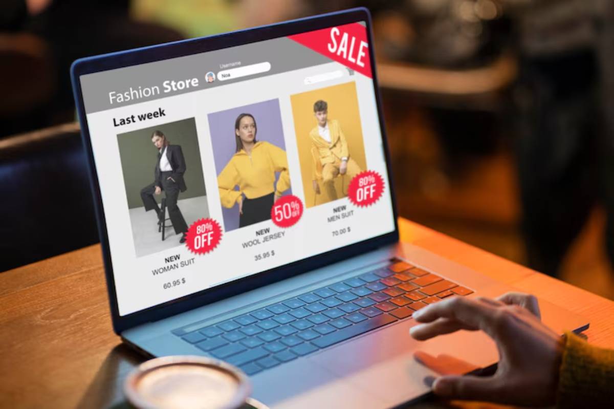 A hand hovers over a laptop displaying a fashion store website with sales on women and men’s clothing, featuring discounts up to 80%. 