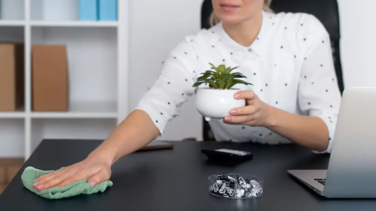 A woman wipes a desk with a cloth while holding a potted plant, surrounded by office supplies and a laptop. 