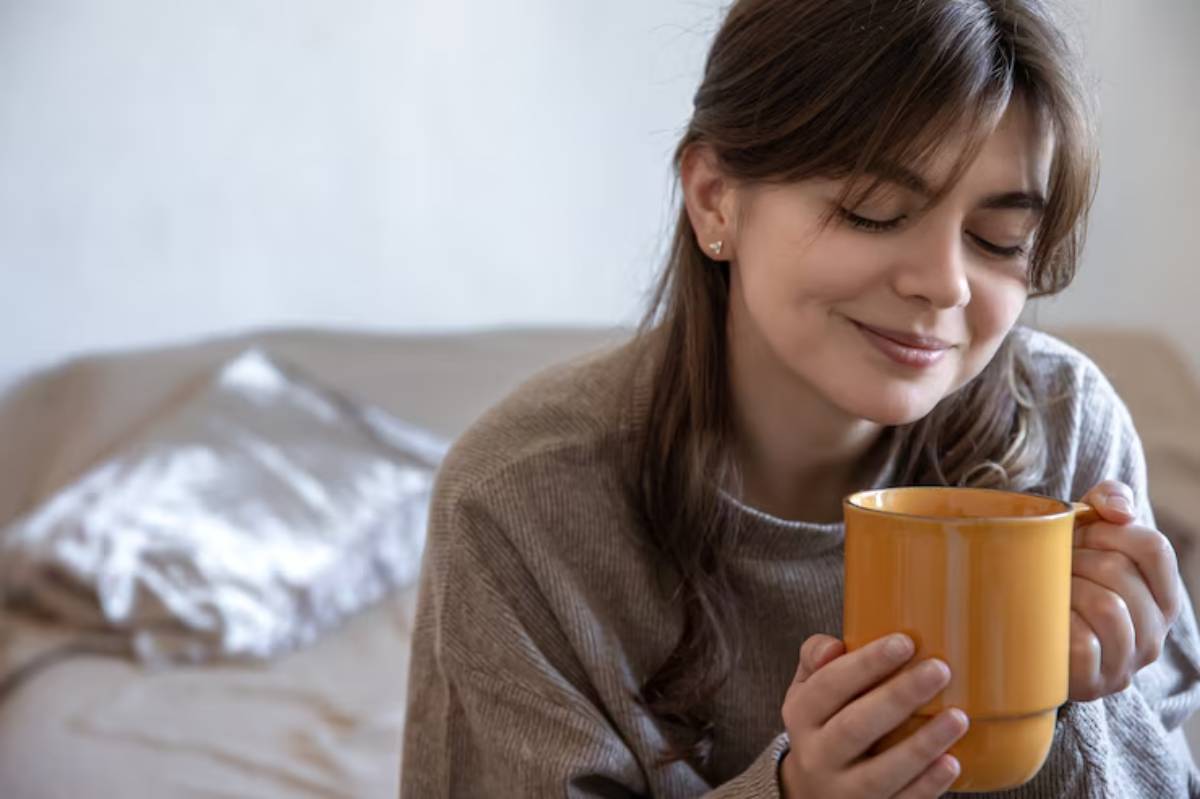 A person with long hair holds a bright orange mug, sitting on a beige couch with a decorative silver pillow in the background. 