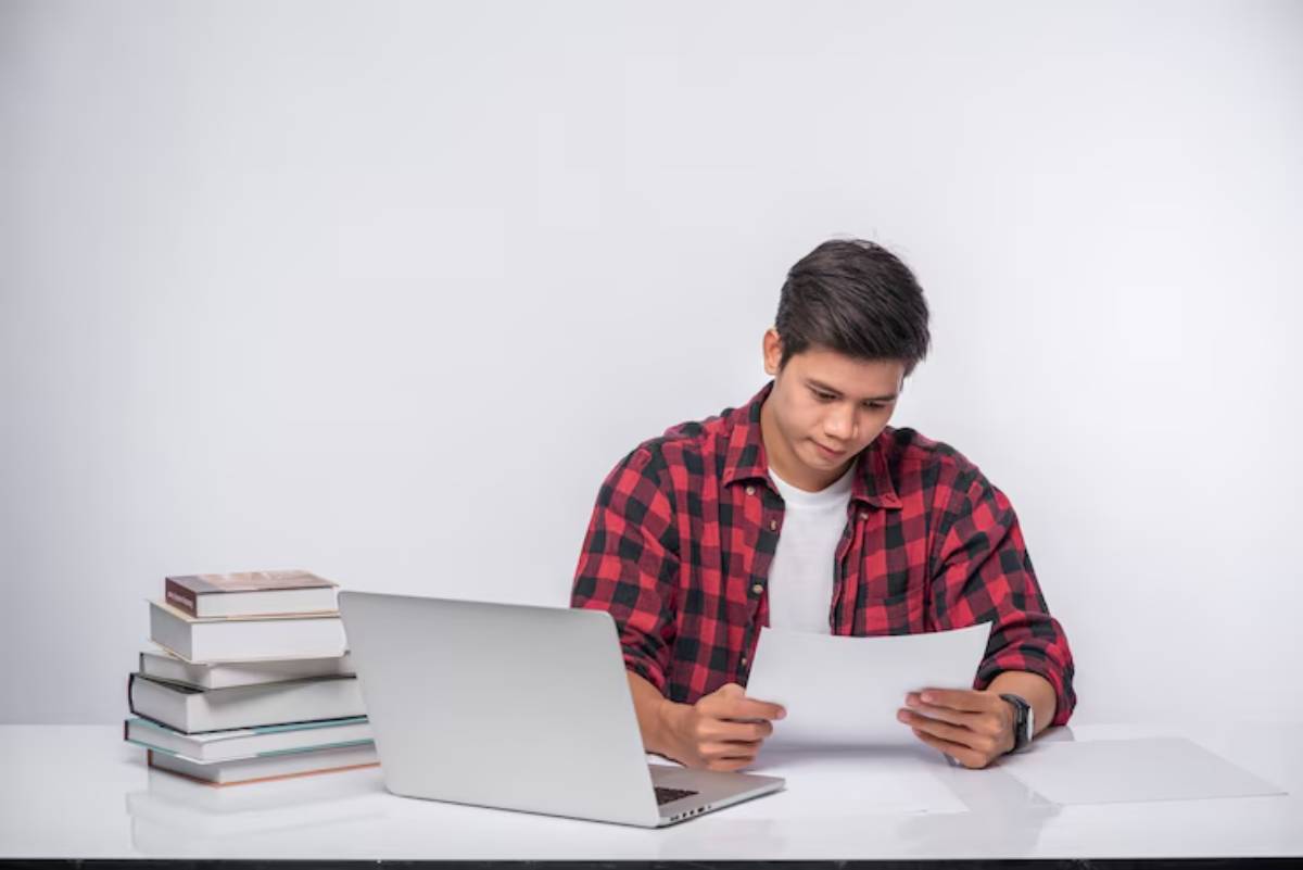 A person in a plaid shirt sits at a table, studying a sheet of paper, with a laptop and stacks of books nearby. 