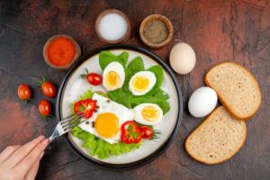 A nutritious breakfast plate with fried egg, boiled eggs, cherry tomatoes, lettuce, and bell peppers, surrounded by spices and bread.