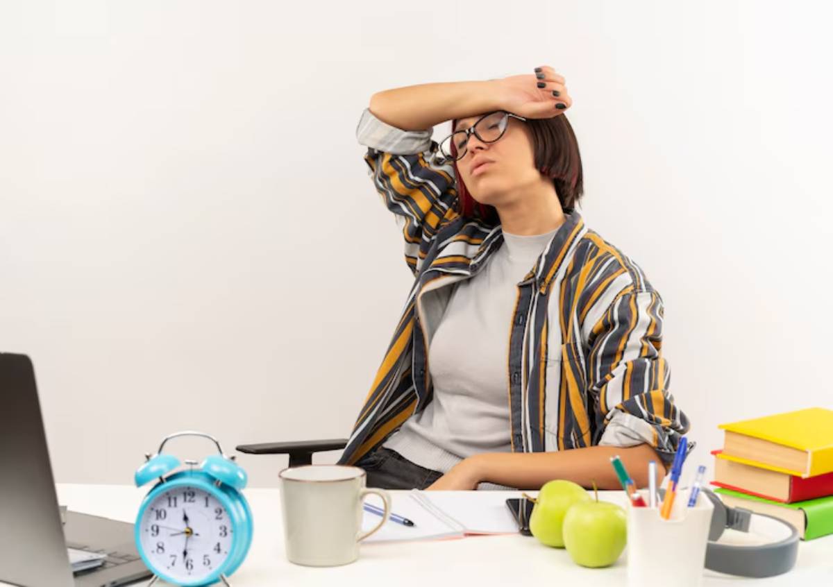 A young person wearing a striped shirt sits at a desk with an alarm clock, a cup, green apples, and colorful notebooks. 