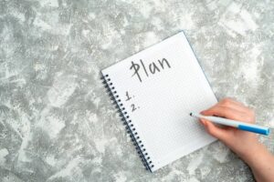 A hand writes Plan with numbered bullet points in a spiral-bound notebook on a textured gray surface.