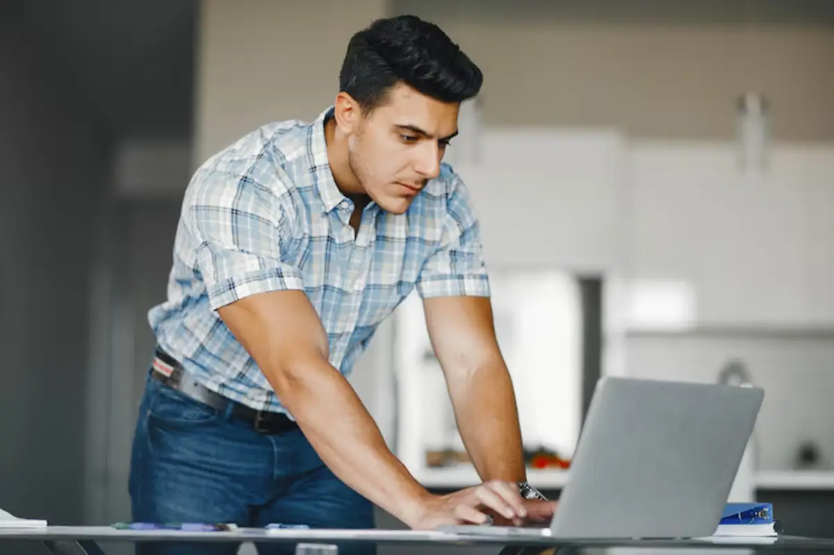 A man in a plaid shirt leans over a laptop on a glass table, engaged in work in a modern kitchen setting. 